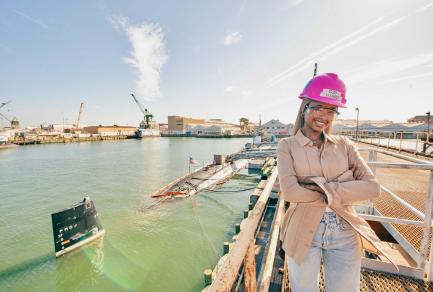 A woman in a pink hard hat posing by a harbor