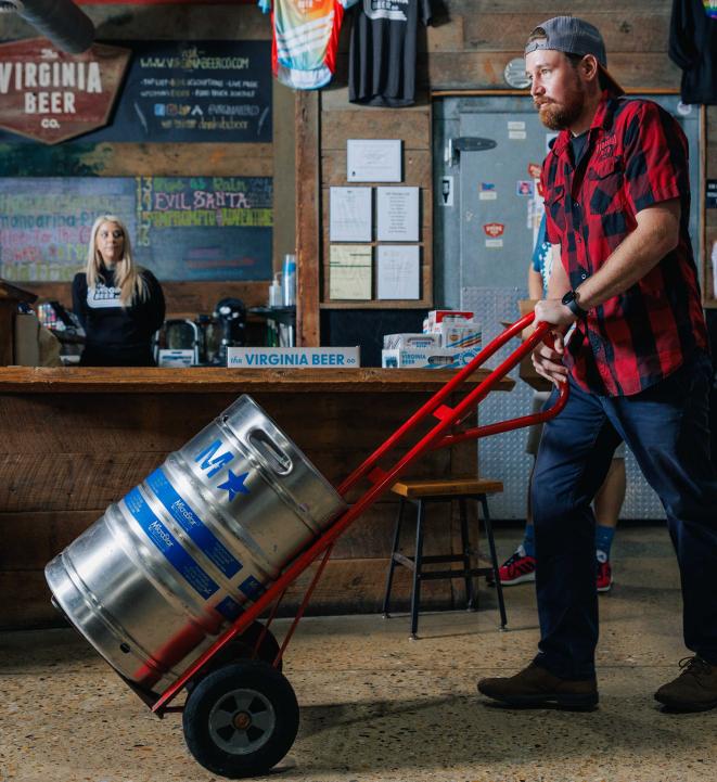 A man wheeling a keg of beer into a bar with a woman in the background