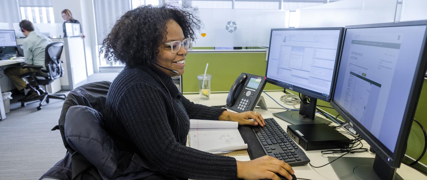 A Black woman talking on the phone and typing on a computer with dual monitors