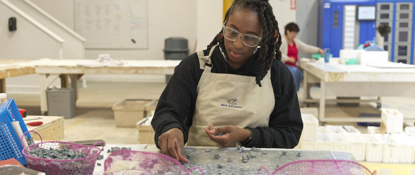 A woman wearing glasses, sitting at a table and using tiles to make a mosaic