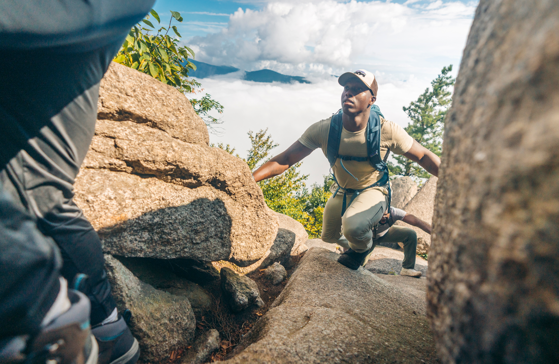 Hikers climbing over large rocks with mountains and clouds in the background.